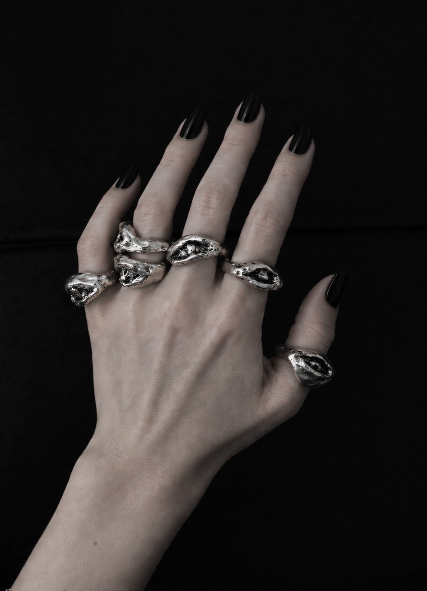 Hand wearing multiple silver rings with black nail polish on a dark background